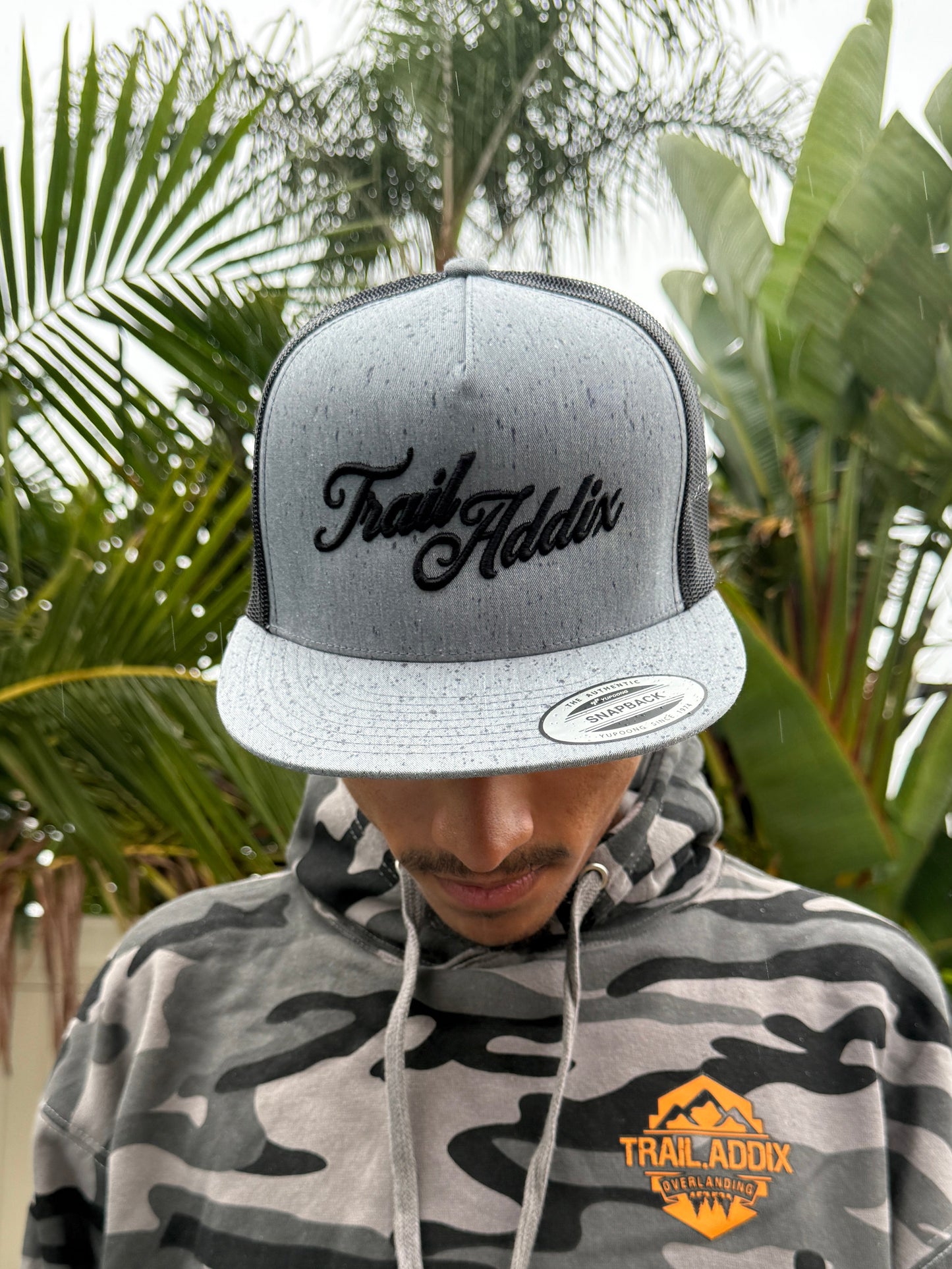 Person wearing a camouflage hoodie and cap with text, standing outdoors with greenery.