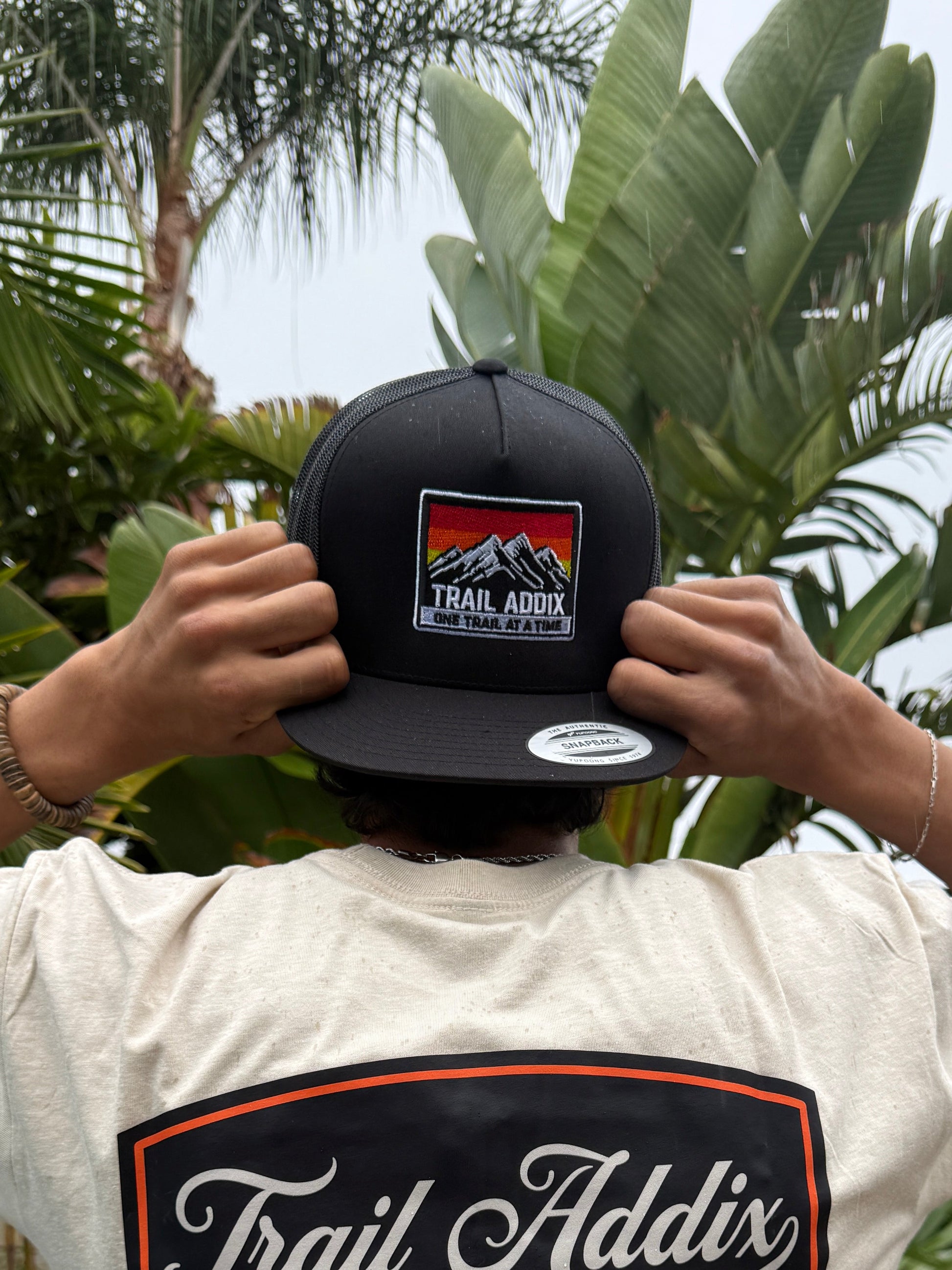 Person wearing a white shirt with a 'Trail Addict' logo, holding a black cap with a similar logo, outdoors.