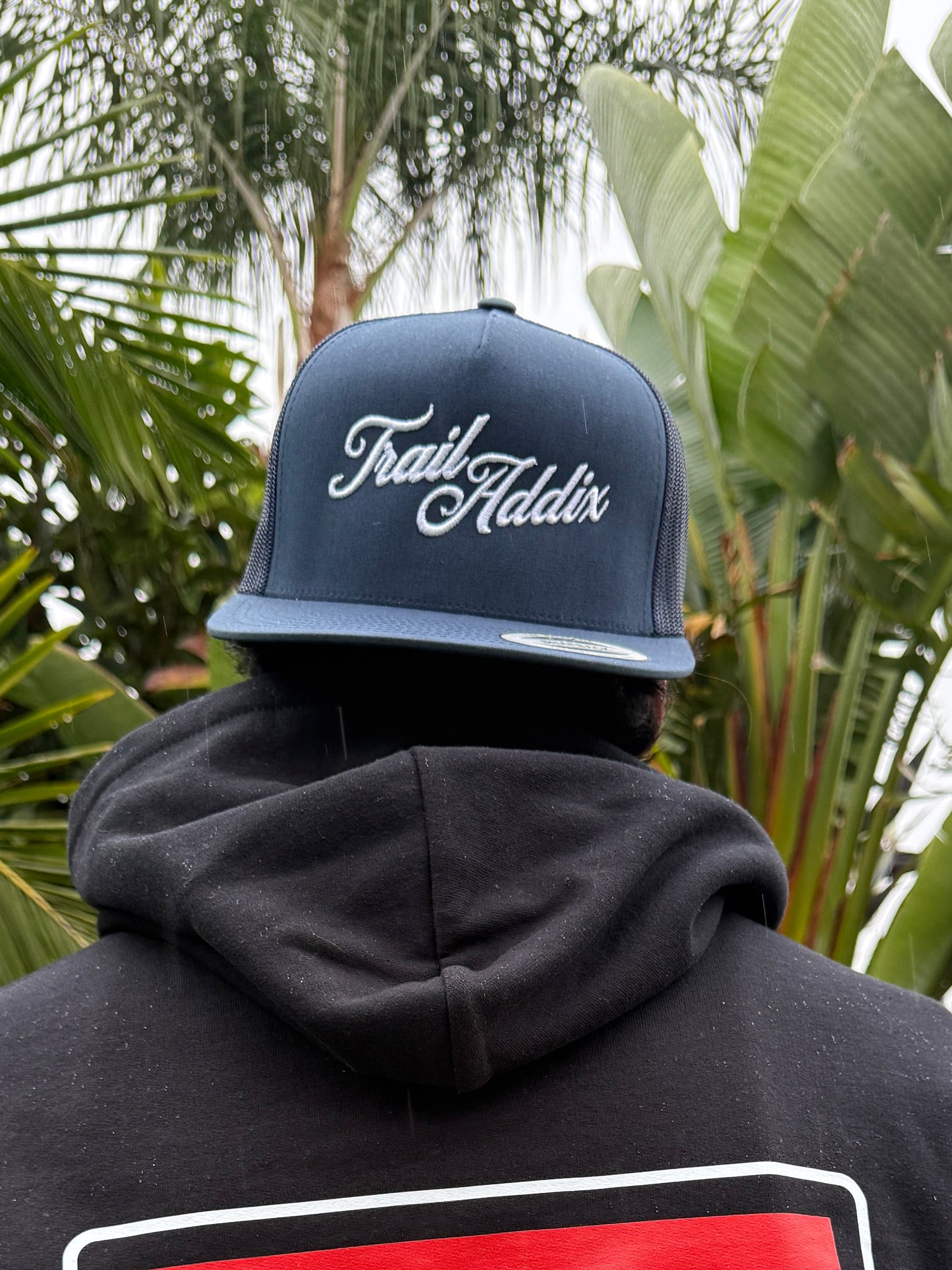 Person wearing a black hoodie with a 'Trail Addix' logo, standing outdoors with plants in the background.